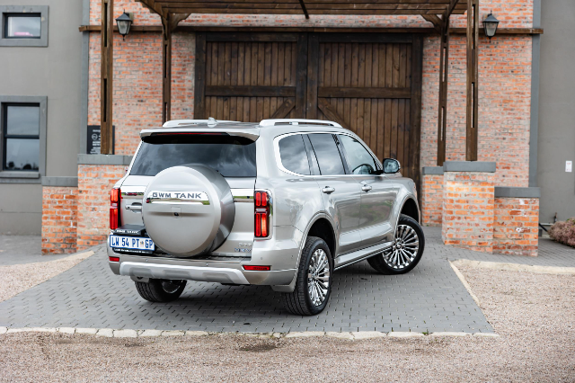 A silver GWM Tank 500 SUV is parked on a paved driveway in front of a rustic brick building with wooden doors. The vehicle's rear features distinctive vertical taillights and a spare tire with the "GWM Tank" logo. The surroundings include a mix of brickwork and greenery.