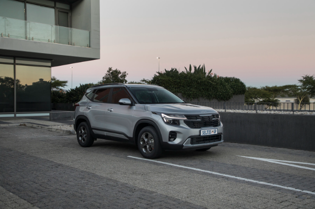 A sleek silver Kia Seltos parked on a paved street beside a modern building with large glass windows. The background features lush green shrubs and a soft evening sky.