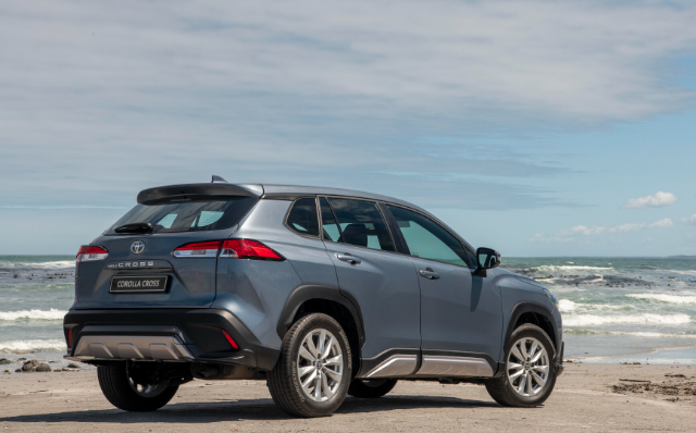A gray Toyota Corolla Cross SUV is parked on a sandy beach with the ocean and waves in the background. The vehicle is viewed from the rear side, showcasing its modern design and sleek lines under a partly cloudy sky.