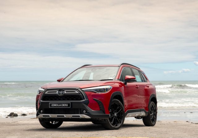 A red Toyota Corolla Cross SUV is parked on a beach with the ocean and a cloudy sky in the background. The vehicle features a prominent front grille, sleek headlights, and black alloy wheels. The badge with "Corolla Cross" is visible on the grille.