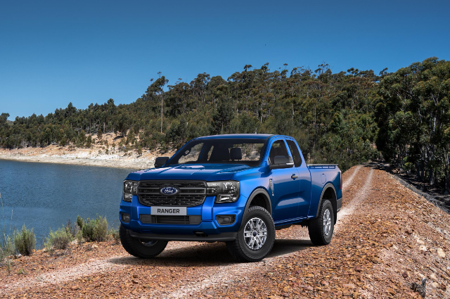 A blue Ford Ranger extended cab truck is parked on a rocky dirt road next to a body of water. In the background, there is a forest of tall trees under a clear blue sky.