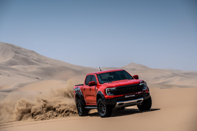 A red Ford Ranger Raptor is driving through a sandy desert landscape, creating a cloud of dust behind it. The background features rolling sand dunes under a clear blue sky.
