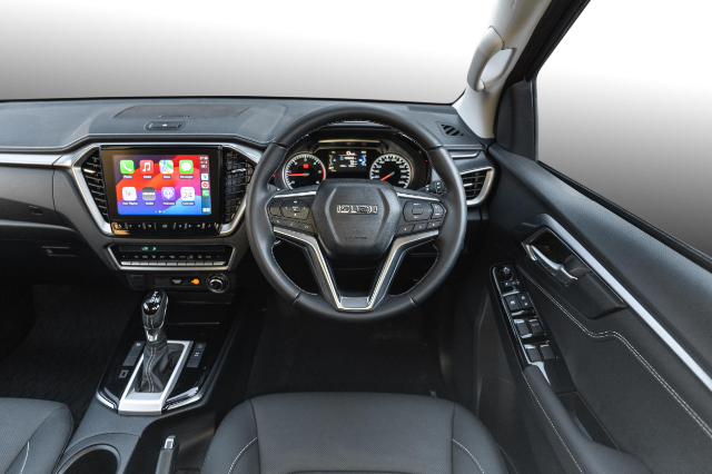 The image shows the interior of an Isuzu D-Max. The dashboard features a central touchscreen infotainment system displaying app icons. The steering wheel, centered in the image, has various control buttons. The gear shift is positioned in the middle console, and the instrument cluster behind the wheel displays speed and other readings. The driver's door panel with window controls is visible on the right. The interior is modern and sleek with a predominantly black color scheme.