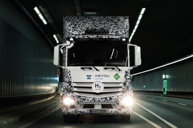 A hydrogen-powered truck with "KEYOU inside" and "H2 Gas" logos is driving on a country road. The truck has a patterned design and the sun is setting behind it, casting a warm glow over a grassy landscape with a pathway and trees in the background.