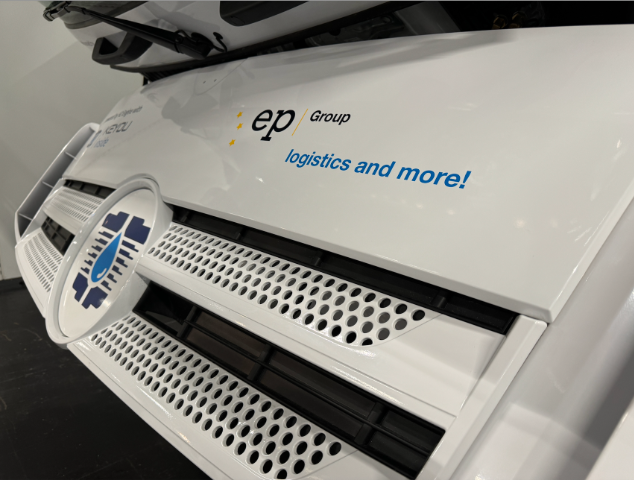 The image shows a close-up of a white vehicle with a logo and text on the front. The logo is circular with a blue and white design. The text next to the logo reads "ep Group logistics and more!" in black and blue letters. The vehicle has a perforated grille and appears to be part of a logistics or transport company.