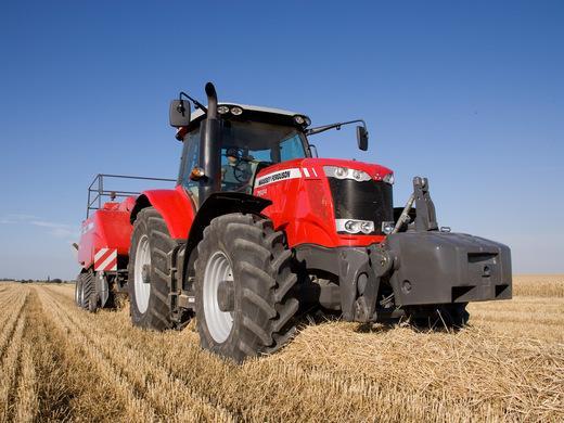 A red Massey Ferguson 7624 Dyna-VT tractor is seen operating in a field of harvested crops. The tractor is equipped with large black tires and is towing a red baler in the background, preparing hay or straw bales. The scene is set under a clear blue sky with golden straw covering the ground.