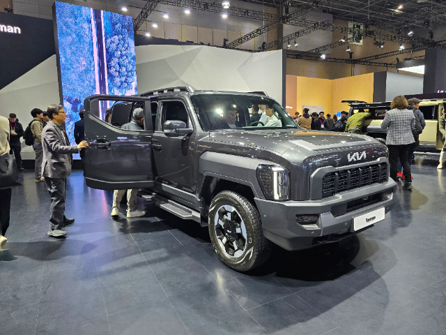 A gray SUV with a bold, rugged design displayed at an indoor automotive event. People are gathered around, with some inspecting the vehicle up close. The SUV features the Kia logo on the grille and has an open front passenger door. A large screen in the background shows an aerial image of a forest and a road, adding to the event's visual ambiance.