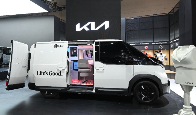 A sleek white Kia electric van is displayed at an indoor exhibition booth. The van features open sliding and rear doors, showcasing a modern interior setup with a rotating chair, illuminated floor, and digital screens. The van displays "Life's Good" branding alongside the LG logo. A large Kia logo is illuminated on the wall in the background, and nearby exhibits, such as merchandise and display stands, surround the area.