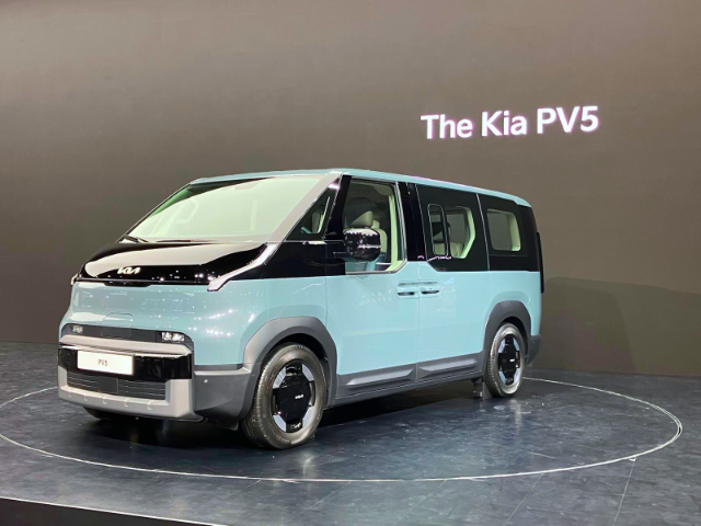 Image of a modern Kia PV5 electric van displayed on a showroom stage. The van has a sleek, futuristic design with a two-tone color scheme of light blue and black. The front grille is minimal, featuring a Kia logo, and the windows have a distinct angular styling. The background is minimalistic with 'The Kia PV5' text projected on a plain wall."