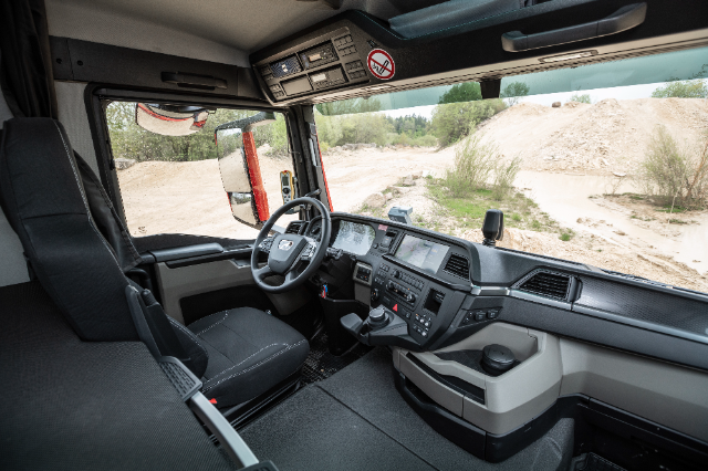 The image shows the interior of a modern truck cabin, designed with ergonomic and functional features. The dashboard incorporates a sleek design with multiple control panels, digital displays, and an infotainment system. The steering wheel has integrated controls, and the driver's seat is spacious and cushioned. A "no smoking" sign is displayed above the windshield. The large side window and windshield provide a clear view of an outdoor landscape with sandy terrain and greenery.
