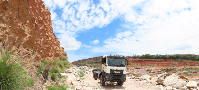 A rugged landscape featuring a white truck on a rocky dirt road. To the left, there is a steep orange rock face with patches of green vegetation. The background shows layers of red soil and greenery under a partly cloudy blue sky.