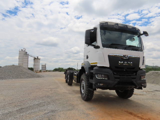 A white heavy-duty truck with the "MAN" logo on the front is parked on a gravel area under a partly cloudy sky. In the background, there are mounds of gravel and industrial silos, suggesting a construction or industrial site.