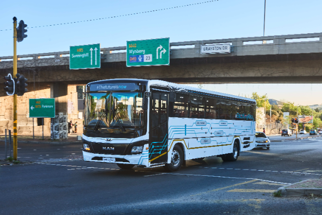 A white electric bus labeled "MAN" with modern, circuit-like blue and yellow graphic designs is driving on a city street. The front display reads "#TheFutureIsNow," and the side incorporates the hashtag "#ElectrifyAfrica." The bus is passing an urban area with visible road signs ("M85 Sunninghill," "M40 Wynberg") on an overpass above, and the setting is bright and sunny with minimal traffic.