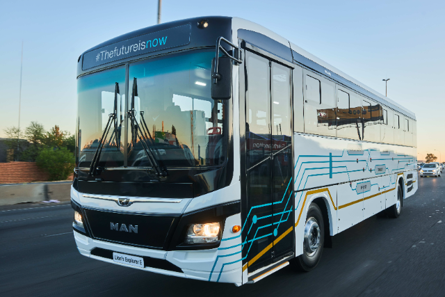 A modern electric bus (MAN Lion's Explorer E) is pictured on a roadway. Its front design is sleek and aerodynamic with a clean white body, black accents, and digital blue graphics along its side, resembling electronic circuits. The destination sign on top reads "#Thefutureisnow." The bus is moving, with its headlights illuminated during daylight. Surrounding background includes other vehicles and an urban roadside setting under a clear sky.