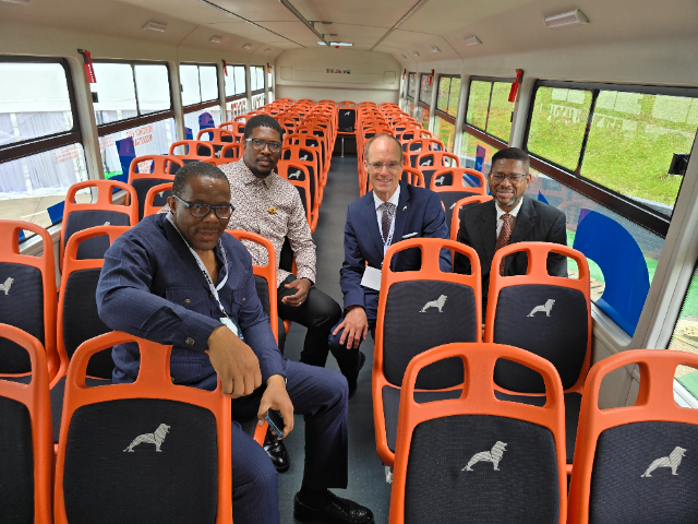 bus with orange seats and a minimalist lion emblem on the seatbacks. The bus features large, clear windows, allowing visibility of the green grass outside. The men, dressed in formal and business casual attire, are smiling and engaged in conversation. The interior of the bus looks spacious and well-lit.