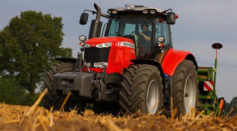 A red Massey Ferguson 7624 Dyna-VT tractor is pictured from a low angle in a field with straw-covered ground. The tractor has large tires and is pulling farming equipment in the background. A green tree and cloudy sky are visible in the distance