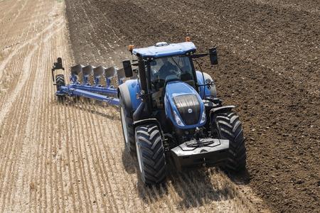A blue New Holland tractor pulling a semi-mounted variable reversible plough on a field. The field is partially ploughed, with one side showing freshly turned soil and the other side showing unploughed stubble. The tractor is in operation, preparing the soil for cultivation.