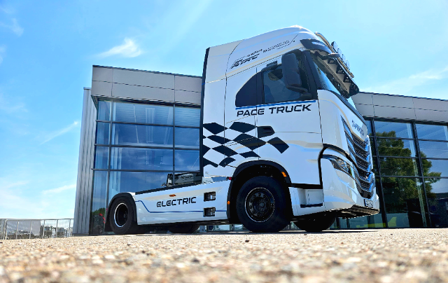 A white electric pace truck with "PACE TRUCK" and "ELECTRIC" written on its side, featuring a checkered flag decal. The truck is positioned in front of a modern glass building under a clear blue sky.