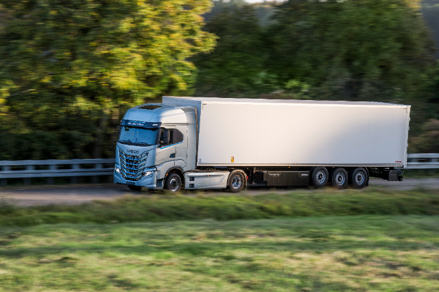 A sleek silver electric truck with "IVECO" branding is shown pulling a white trailer on a road surrounded by greenery. The truck is in motion, with blurred vegetation in the background, indicating speed. The scene is lit by natural daylight.