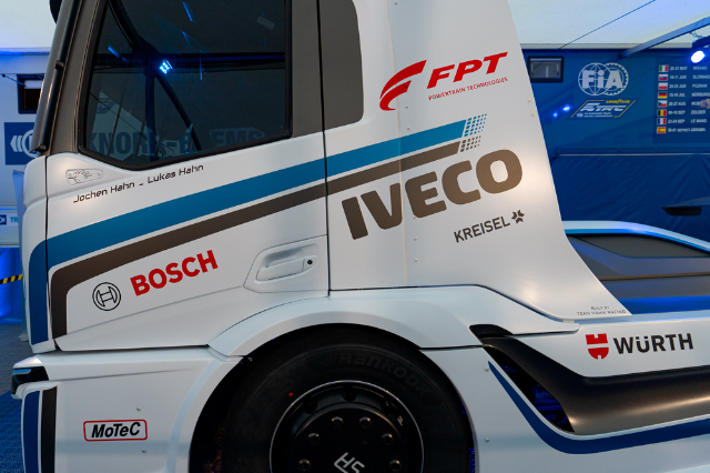 A close-up side view of a white race truck covered with logos of various sponsors. The words “Iveco” and “Bosch” are prominently displayed alongside other logos like “FPT Powertrain Technologies,” “Kreisel,” “Würth,” and “MoTeC.” The names “Jochen Hahn” and “Lukas Hahn” are visible near the cabin door. The truck is parked in an indoor environment with a blue-lit background, and part of the tire and body design is visible.