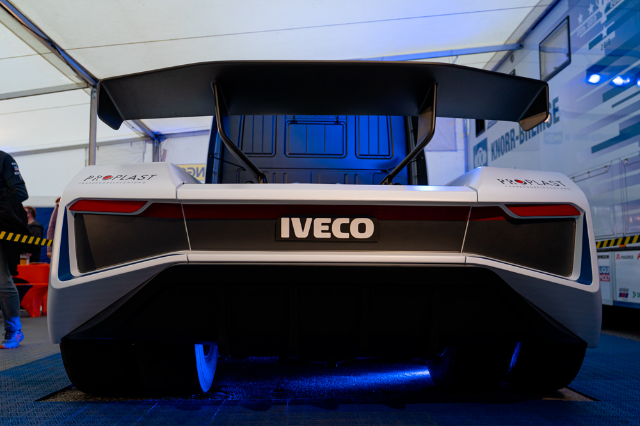 A rear view of a sleek white race truck with the "IVECO" logo prominently displayed in the center. The vehicle features sharp, angular taillights and an oversized black spoiler. It is in a well-lit indoor setting, with blue undertones reflected underneath the vehicle. The background shows a tent-like structure and partial views of other vehicles and equipment