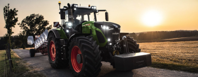 A green Fendt 936 Vario tractor is driving down a rural road at sunset. The tractor is equipped with large tires, bright headlights, and is pulling agricultural equipment. The background features open fields, a tree, and a warm orange glow from the setting sun.