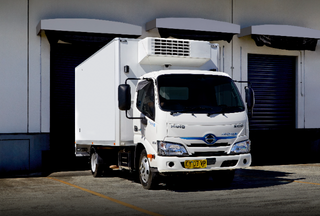 A white Hino 300 series hybrid refrigerated truck is parked outside a warehouse, positioned in front of a loading dock with closed roller doors. The vehicle features a refrigeration unit on top of its cargo area and a yellow license plate on the front. The truck is illuminated by sunlight, highlighting its clean and modern design. The background includes concrete pavement and the white walls of the industrial building.
