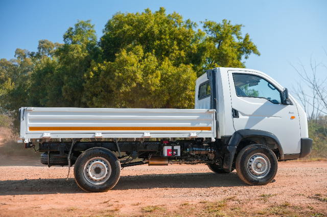 A compact white flatbed truck is parked on a dirt path. The vehicle has a simple, streamlined design with a white cab and a long, open cargo bed with orange reflective strips. Its wheels are clean, and the background shows lush green trees under a clear blue sky.