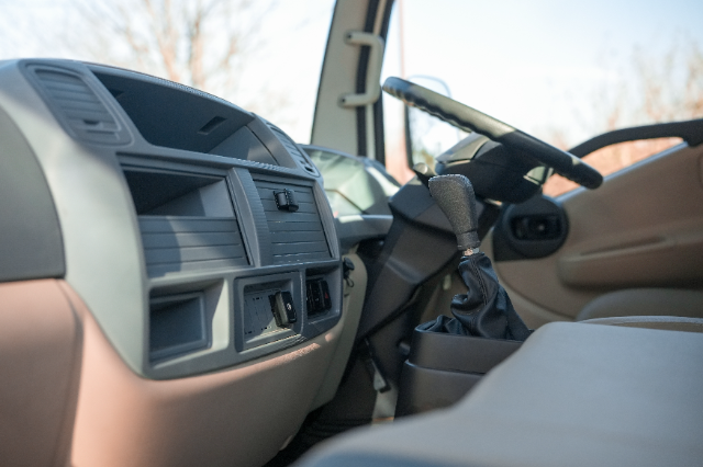 A close-up view of a vehicle's interior focusing on the dashboard and steering area. The dashboard includes uncluttered compartments and smooth paneling, while the gear stick with a fabric cover is visible in the foreground. The steering wheel and part of the driver's side door are visible in the background. Natural light enters the vehicle, and blurred tree branches can be seen through the windows.