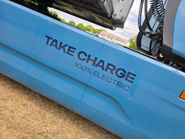 A close-up angle of the side of a light blue vehicle with "TAKE CHARGE 100% ELECTRIC" prominently displayed in bold text on its body. In the background, there is an open component or machinery at the top, with visible coiled wires, and blurry greenery along with a building visible in the distance. The ground beneath the vehicle appears to be dry grass.