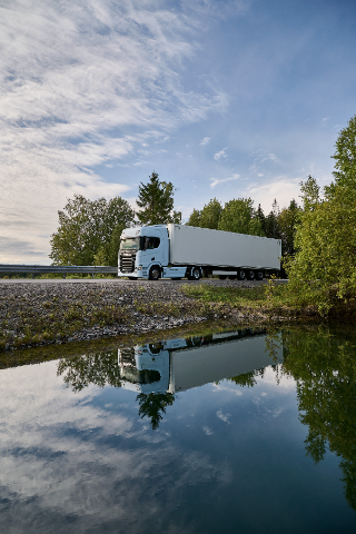 A white semi-truck with a trailer is driving on a paved road surrounded by green trees and shrubs. The truck's reflection is visible in a calm body of water below, while the sky above is partly cloudy and blue, contributing to a serene and picturesque scene.