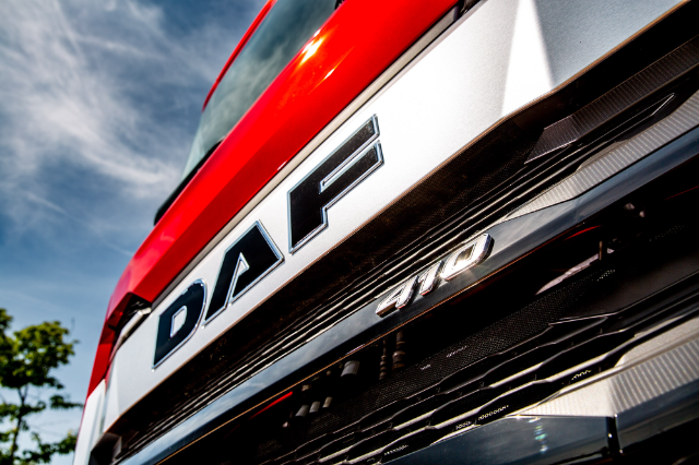 Close-up view of the front grille of a DAF truck, featuring the bold "DAF" logo prominently displayed on a metallic background. Below, the number "410" is visible, indicating a model or specification. The truck has a red exterior, with a blue sky and some tree leaves blurred in the background, adding a dynamic and outdoor setting.