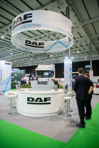  A trade show booth for DAF prominently featuring the company's logo and the slogan "Powering Your Success" on a large circular hanging banner above. The booth includes a white rounded counter with stools, brochures, and a sleek modern design. A large DAF truck is displayed in the background, surrounded by other exhibition materials and attendees engaging in discussions. The floor is partly covered with green carpeting, and the exhibition hall has a high industrial-style ceiling.