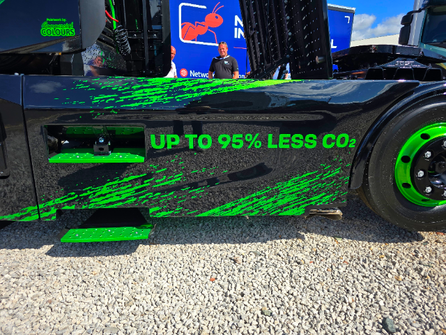 A close-up image of the side of a black semi-truck or heavy vehicle with green accents and text. The text on the vehicle reads, "UP TO 95% LESS CO₂" in bold green lettering. The design features a green streak pattern along the side. The wheel has a black rim with green highlights. The background reveals a gravel floor, part of a blue sign with a red ant logo and text, and two individuals standing near the truck.
