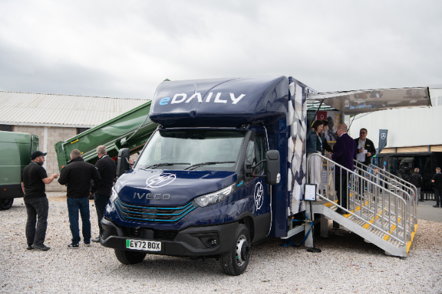 A navy blue Iveco eDaily electric vehicle is on display outdoors at an event. The van has "eDAILY" branding on the front with a lightning bolt symbol on the hood. A set of stairs leads to an open side door with a canopy, where people are gathered inside and outside the vehicle, engaging in discussions. The background includes another green vehicle and a warehouse-type building under a cloudy sky.