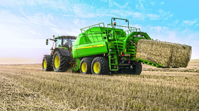 A bright green agricultural machine attached to a tractor is working in a harvested field under a blue, slightly cloudy sky. The machine, likely a baler, is ejecting a large, rectangular hay bale. The field is filled with short stubble of cut crops, and the machinery’s large yellow tires are visible.