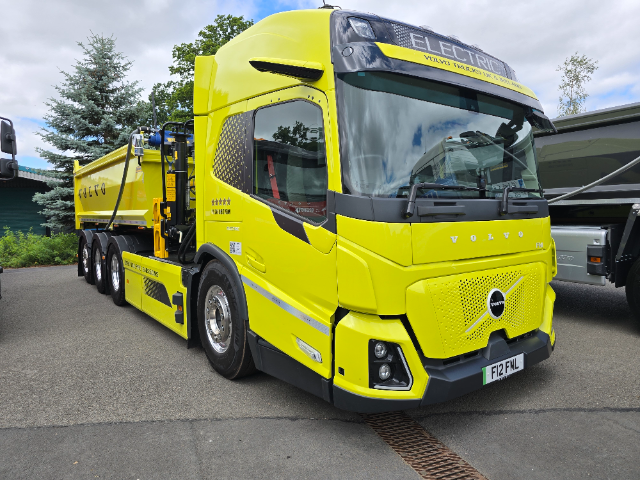 A bright yellow Volvo electric truck is shown in a parked position. The truck features a modern, aerodynamic design and displays the text "ELECTRIC" on the front windscreen. The side of the truck's trailer has the Volvo logo and additional markings about emissions, indicating it is focused on sustainability. The vehicle is situated on a paved area with some greenery and trees in the background under a cloudy sky.