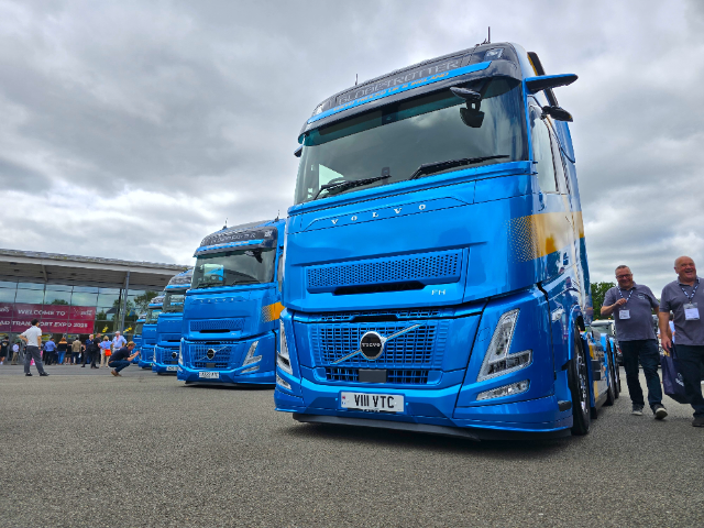 A close-up view of two bright blue Volvo FH trucks parked outdoors at a transport expo event. The trucks are clean and prominently display their logos and design details. A group of people is gathered in the background near a building with signage that reads "Welcome to Road Transport Expo 2025." Two men are walking and smiling on the right side of the image under a cloudy sky.