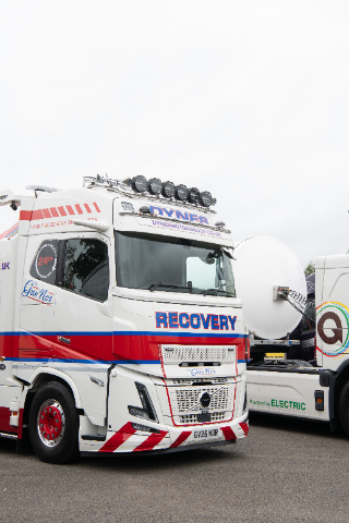 A large white recovery truck with red and blue accents is prominently featured in the image. The truck has "RECOVERY" written in bold letters on the front grille and "DYNES" displayed above the windshield, along with additional details like "Heavy Recovery Specialists" along its side. The truck is equipped with a set of roof lights, and its license plate reads "GV25 NOR." Another vehicle with "Powered by Electric" signage is partially visible in the background. The setting appears to be an outdoor parking or display area under a cloudy sky.