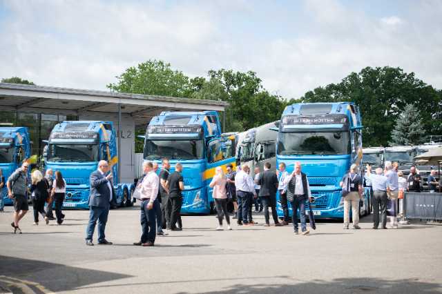 A group of people gathered around a display of blue Volvo Globetrotter trucks in an outdoor exhibition setting. The trucks are parked in front of a building labeled "Hall 2." The attendees, dressed in a mix of business and casual attire, are walking, conversing, and observing the vehicles. The scene is set on a bright, partly sunny day with greenery and trees visible in the background.