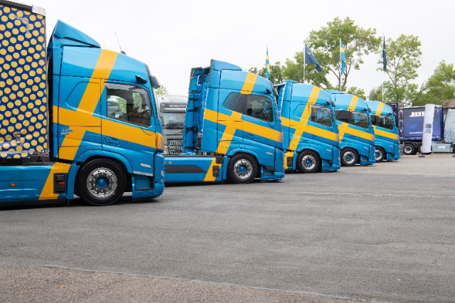 A lineup of bright blue semi-trucks featuring bold yellow diagonal stripes reminiscent of the Swedish flag. The trucks are parked in a row on a paved lot, with trees, flags, and other trucks visible in the background. The truck on the far left has a polka-dot pattern on its trailer.