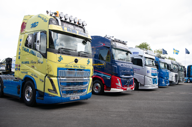 A row of colorful, well-maintained Volvo trucks on display in an outdoor setting. The first truck in the foreground is painted yellow and blue, with visible branding for "T. Alun Jones Ltd." Subsequent trucks feature various designs and colors, including blue, red, and silver. The background shows trees, flags, and a partly cloudy sky. The setting resembles a truck exhibition or gathering.