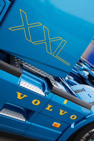  Close-up view of a blue Volvo truck featuring the "XXL" text in bold yellow letters on the side of the vehicle's upper portion. The lower section displays the "VOLVO" logo in matching yellow. The image highlights the metallic finish, footstep details, and part of the truck chassis. The background shows another blue vehicle blurred in the distance.